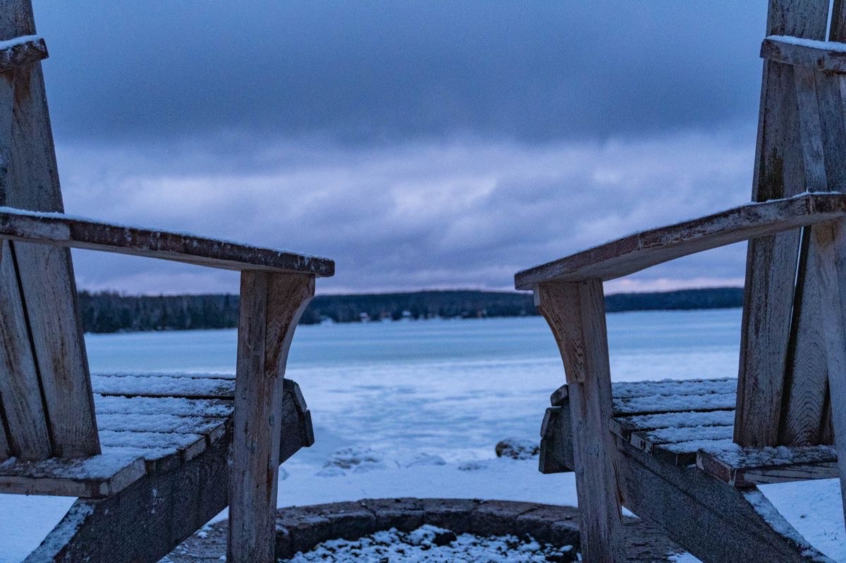 picture of two lawn chairs covered in snow, looking out onto a lake with mountains behind it